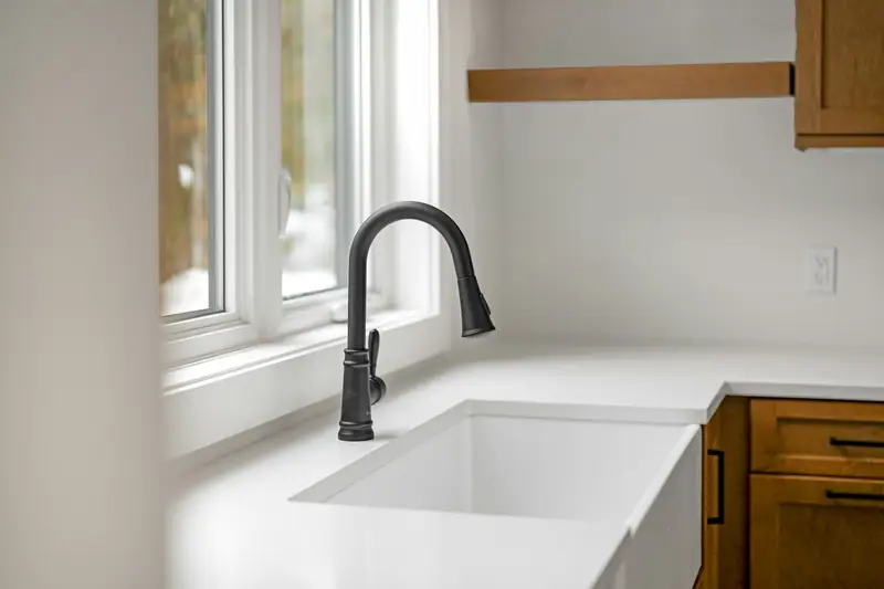 A matte black gooseneck faucet over a white apron-front sink, set below a bright window with light wood cabinets to the right