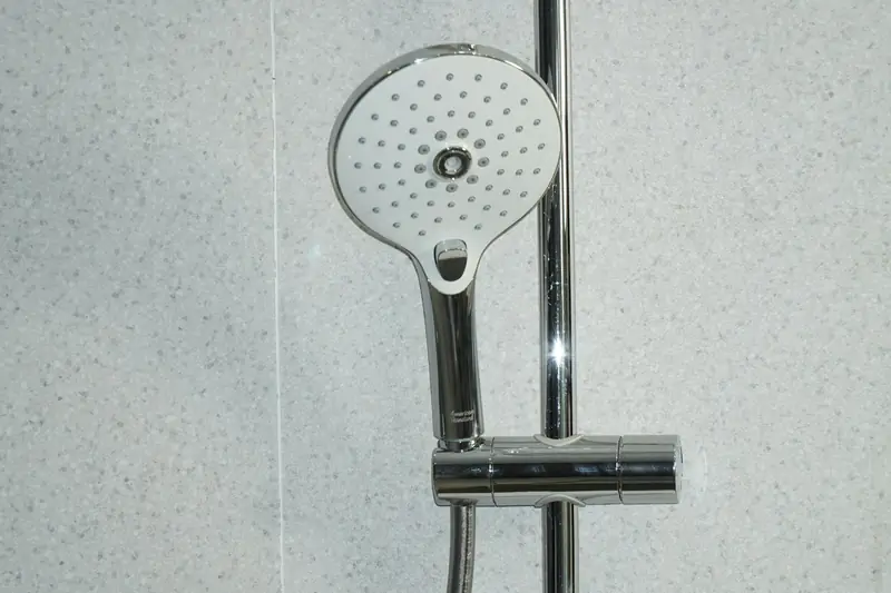 A round chrome hand shower with a white spray face, clipped to a slide bar against a speckled gray wall