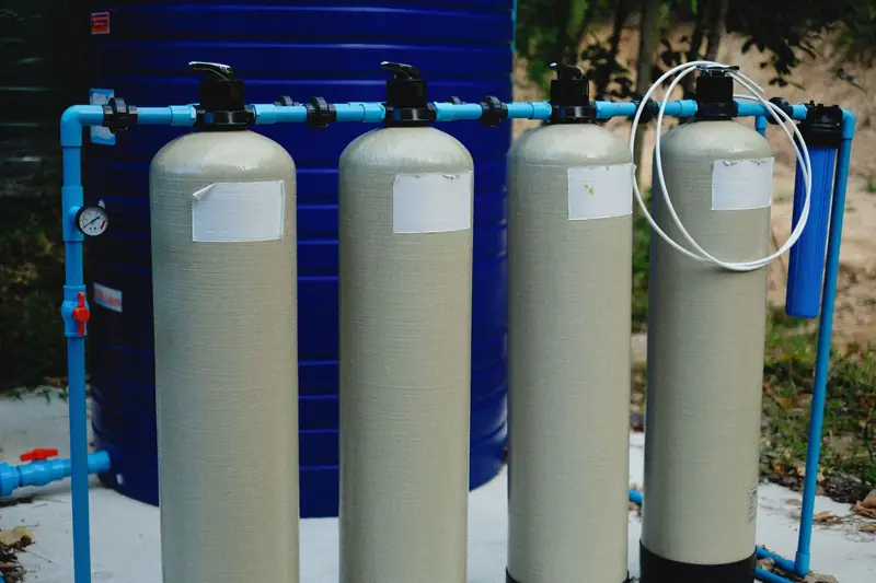 Four beige fiberglass treatment tanks in a row, joined by blue plastic pipe with a small pressure gauge on the left